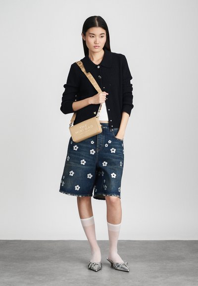 Young woman wearing black knit cardigan, denim shorts with white floral patterns, sheer white socks, silver pointed shoes, and beige shoulder bag.