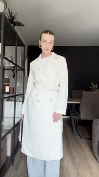 Hvit dobbeltspent trenchcoat med belte i midjen, avsluttet med sølvknapper. Brukt over en lys topp og parret med stripete bukser.