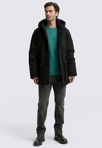 Svart parkas med huva, med dragkedja och knappstängning, texturerat tyg och flera fickor, bärs över en tealfärgad tröja och grå jeans.