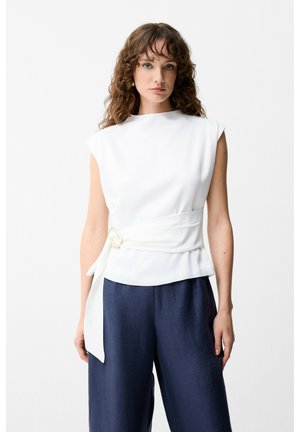 Femme aux cheveux bouclés portant un haut blanc sans manches avec une large ceinture et un pantalon large bleu marine sur fond blanc.