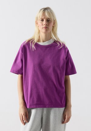 Jonge vrouw met blond haar, gekleed in een losse paarse T-shirt over een wit shirt en grijze joggingbroek, staand tegen een effen achtergrond.