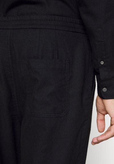 Roupa preta com um bolso lateral e punho abotoado. O material parece suave com uma leve textura. Design funcional e minimalista.