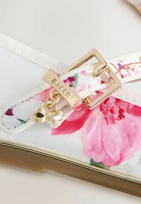 Sandále s kvetinovým vzorom, ktoré majú biely textilný zvršok s ružovými kvetmi, zlatú pracku a ozdobný prívesok v tvare mašle.