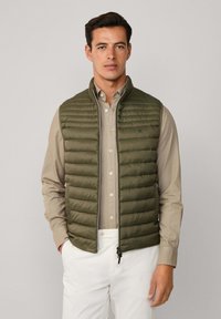 Oliivivihreä pehmustettu liivi, jossa on pystysuuntainen tikkaus, etuvetoketju ja pystykaulus. Sen päällä on vaalea ruskea nappipaita.