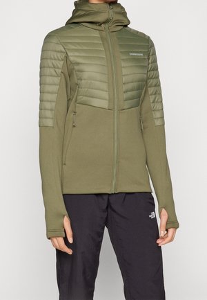 Mujer usando una chaqueta híbrida acolchada y de forro polar en verde oliva con orificios para los pulgares, combinada con pantalones de exterior negros.