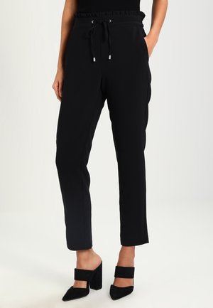 Vrouw draagt zwarte taps toelopende broek met trekkoord in de taille en zwarte hoge hak mules, staand tegen een effen lichte achtergrond.