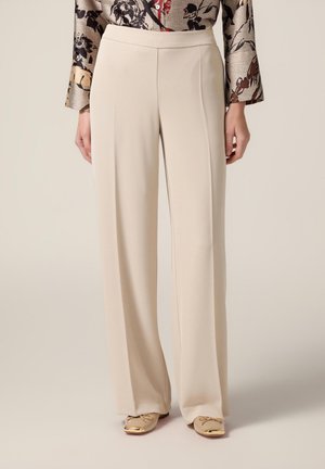 Beige Hochwasserhosen aus glattem Stoff mit hoher Taille und gefaltetem Vorderteil für einen maßgeschneiderten Look. Kombiniert mit hellen Schuhen.