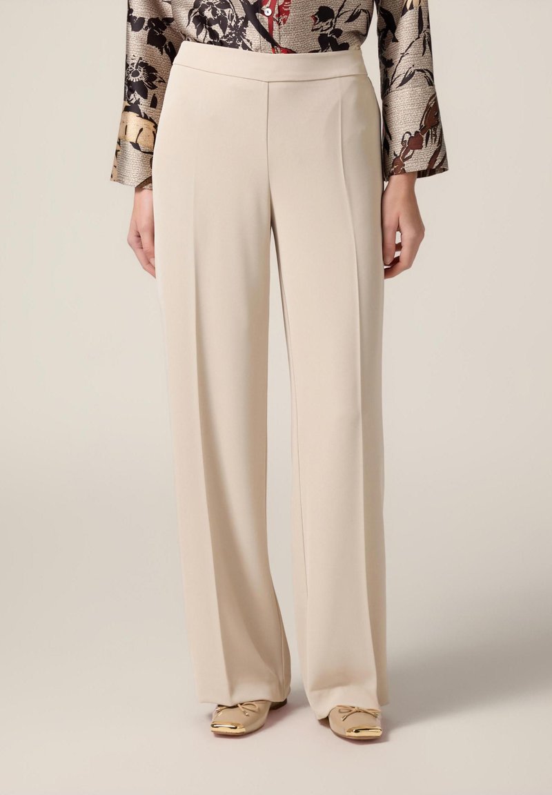 Pantaloni beige a gamba larga realizzati in tessuto morbido, con vita alta e pieghe frontali per un look sartoriale. Abbinati a scarpe di colore chiaro.