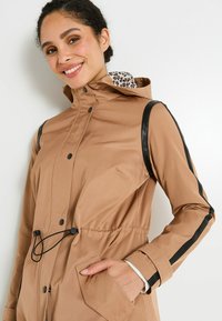 Femme souriante portant une veste à capuche beige avec des finitions noires sur les manches, les mains dans les poches, et un imprimé léopard à l'intérieur de la capuche sur fond blanc.