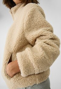Beżowa kurtka z polaru o teksturowanej, puszystej powierzchni; posiada wysoki kołnierz i boczne kieszenie, pokazana w widoku z bliska.