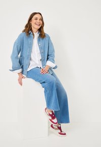 Modèle assis sur un piédestal blanc, portant une veste en denim bleu clair, une chemise blanche et un jean large assorti, complété par des baskets multicolores.