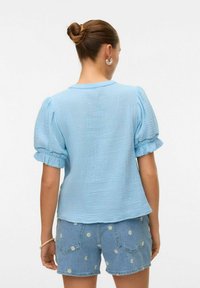 Camisetta azzurro chiaro a maniche corte con tessuto texturizzato, maniche a sbuffo raccolte e scollatura rotonda, abbinata a pantaloni di jeans corti con motivi floreali.
