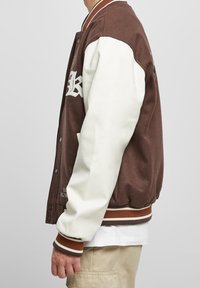 Giacca varsity marrone con maniche in pelle bianca, polsini e colletto a coste. Presenta un logo sul petto e righe a contrasto all'orlo.
