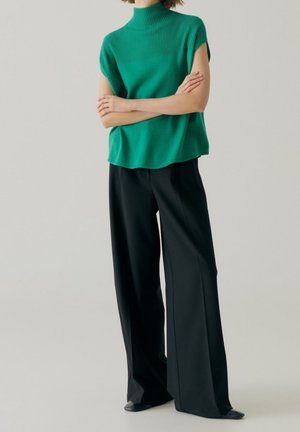 Nainen, joka pukeutunut vihreään, hihattomaan neulepaitaan, jossa on korkea kaulus, ja leveisiin mustiin housuihin, seisoo käsivarret ristissä.