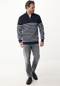 Uomo che indossa un maglione a mezza zip a righe navy e bianche, jeans grigi chiari e stivali neri, in piedi davanti a uno sfondo bianco.