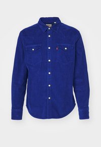 Chemise en velours côtelé bleu à manches longues avec fermeture boutonnée sur le devant et deux poches poitrine boutonnées sur fond blanc uni.