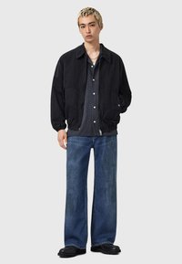 Nuori mies, jolla on lyhyet vaaleat hiukset, pukeutuneena mustaan takkiin, tummaan napilliseen paitaan, leveisiin sinisiin farkkuihin ja mustiin kenkiin, seisoo harmaata taustaa vasten.