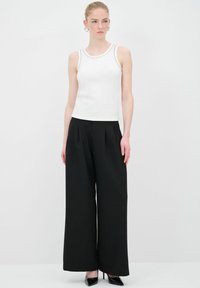 Top bianco senza maniche con borchie decorative sul colletto, abbinato a pantaloni neri a vita alta con gamba larga e tacchi a punta neri.