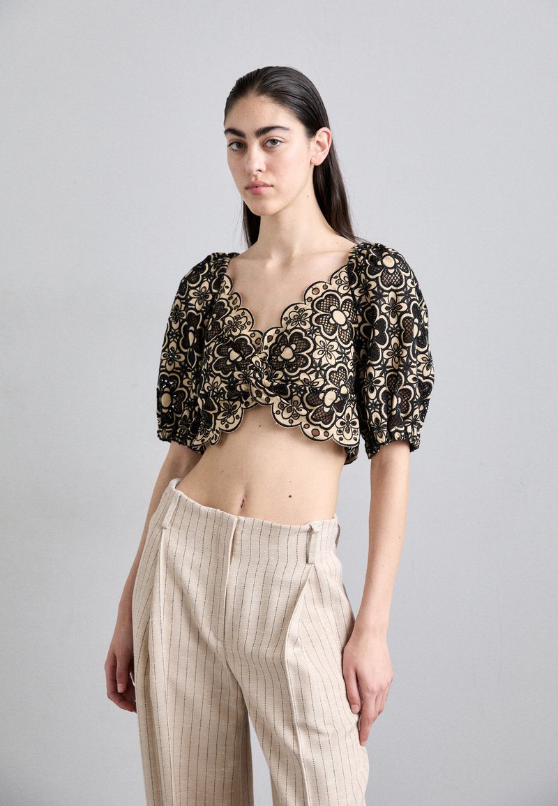 Young woman with long dark hair wearing a black lace floral crop top and high-waisted beige pinstripe pants, standing against a gray background.