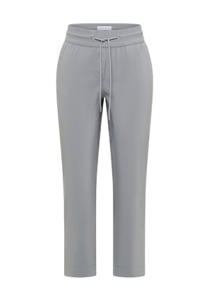 Gros plan d'une personne portant un pantalon gris avec cordon et taille élastique et un t-shirt jaune, main reposant sur le côté.