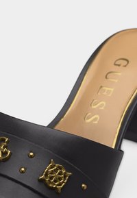 Μαύρο δερμάτινο mule παπούτσι με χρυσά διακοσμητικά καρφιά και μπεζ εσωτερική σόλα με την επωνυμία "GUESS" σε χρυσά γράμματα.