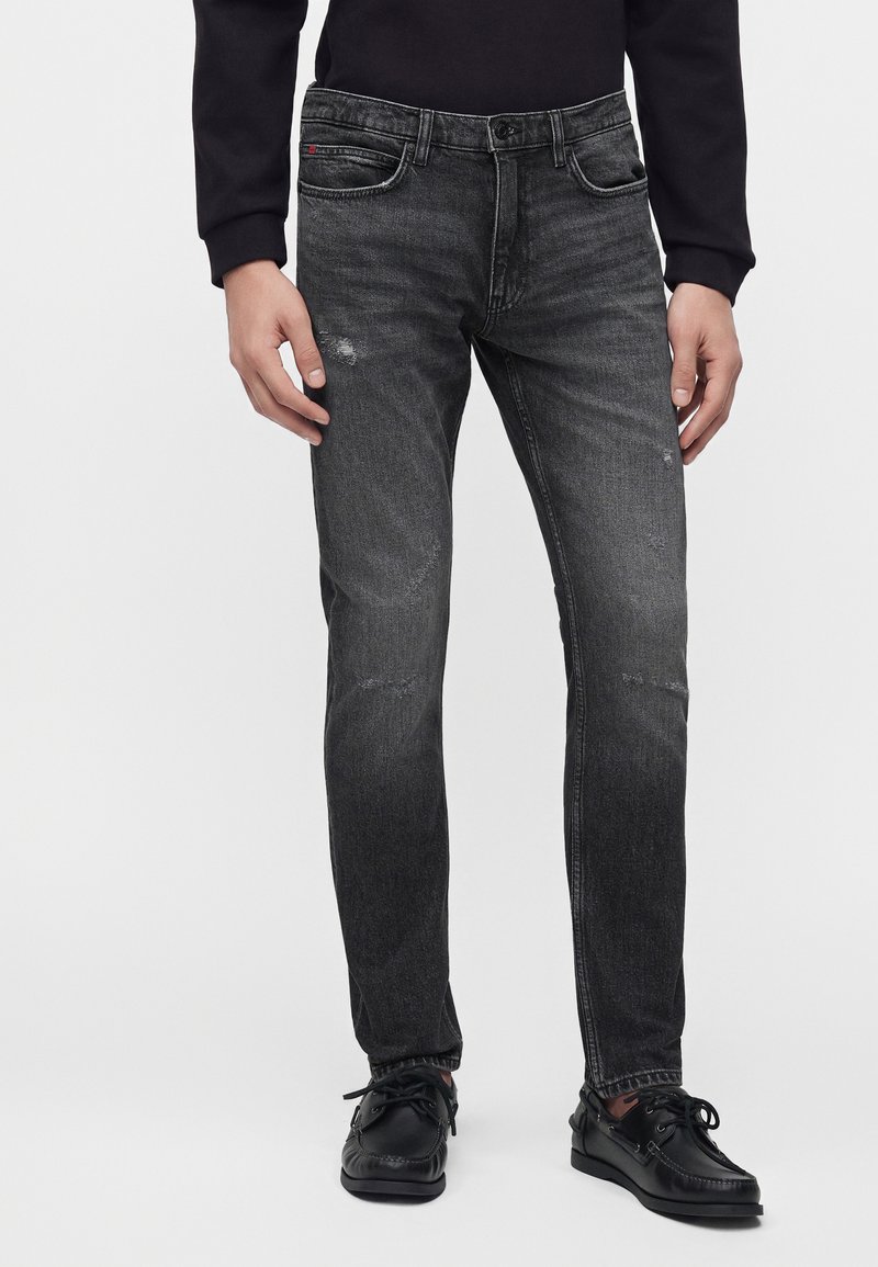 Homme portant un jean slim noir délavé avec de légères déchirures et des chaussures noires à lacets sur un fond uni.