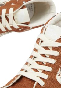 Sneakers in tela marrone con lacci bianchi, caratterizzati da un design a fantasia. L'interno è foderato con un tessuto di colore chiaro. Texture morbida.