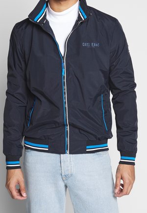 Mann trägt eine dunkelblaue Cars Jeans Jacke mit blau-weiß gestreiften Abschlüssen und hellblauen Jeans vor einem einfarbigen Hintergrund.