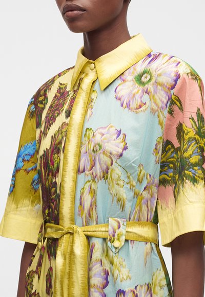 Femme portant une robe fleurie multicolore avec un col jaune, des manches courtes, des boutons à l'avant et une ceinture assortie nouée à la taille.