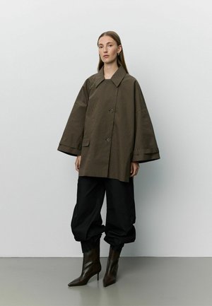 Donna con cappotto marrone a quadri oversize, pantaloni neri larghi infilati negli stivali con tacco marrone scuro, in piedi contro un muro semplice.