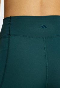 Tamnozelene tajice od glatkog, rastezljivog materijala. Imaju bešavne pojas i suptilan logo na leđima. Detalj ravnog šava.