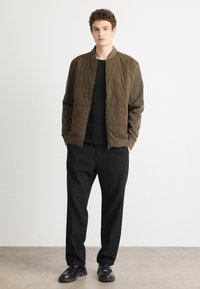Olive green bomber jacket with quilted texture, front zipper, ribbed cuffs, worn over a black sweater and black wide-leg trousers. Black shoes.