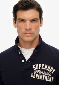Polo de algodón azul marino con cuello texturizado, dos botones y texto bordado en blanco que dice "SUPERDRY Departamento Atlético".