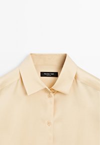 Chemise beige à manches courtes avec un col classique, présentant une texture lisse et une patte de boutonnage. L'étiquette indique "Massimo Dutti".