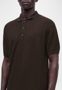 Homme portant un polo en maille texturée marron foncé à manches courtes avec trois boutons sur un fond clair.