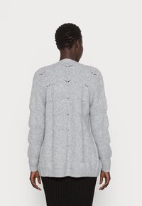 Ljust grå stickad cardigan med texturerade kabelmönster, långa ärmar och en något förlängd bakdel. Inga synliga stängningar eller detaljer.