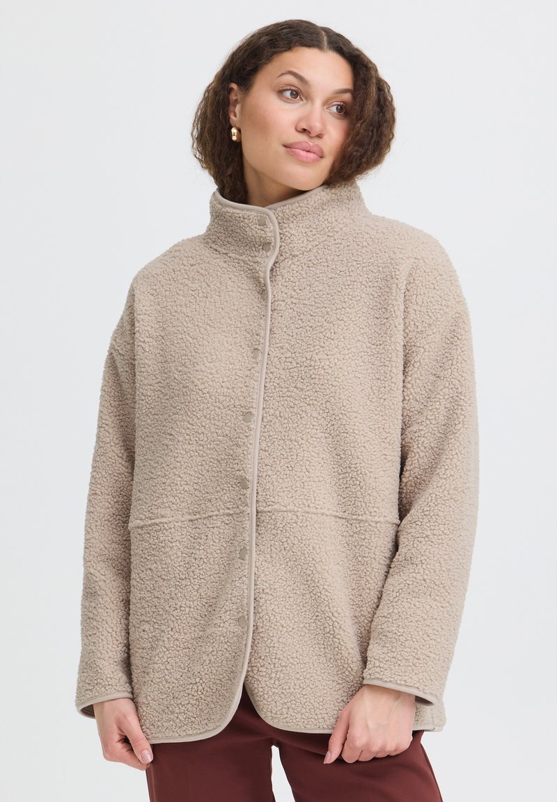 Femme portant une veste sherpa en polaire beige boutonnée avec un col montant et un pantalon marron sur un fond blanc uni.