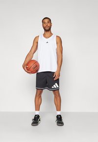 Top de malha branca com detalhes pretos, combinado com calções pretos com riscas brancas e um logo. Segurando uma bola de basquete. Ténis de desporto pretos.