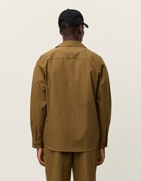 Barna gombos ing, laza szabással, hosszú ujjakkal és gallérral. Simább textúrával és lekerekített aljjal rendelkezik. Fekete sapka, amit hátrafelé viselnek.