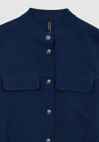 Camicia blu navy con colletto alla cinese, chiusura con bottoni argento e due tasche frontali, realizzata in tessuto liscio con una trama uniforme.