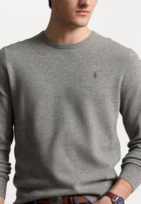 Pull gris en maille avec un col rond et des poignets côtelés, orné d'un petit logo brodé sur la poitrine. Porté avec une ceinture marron.