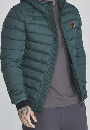 Homme portant une veste matelassée vert foncé par-dessus un t-shirt gris et un pantalon foncé, avec des tatouages visibles sur la main gauche et le cou.