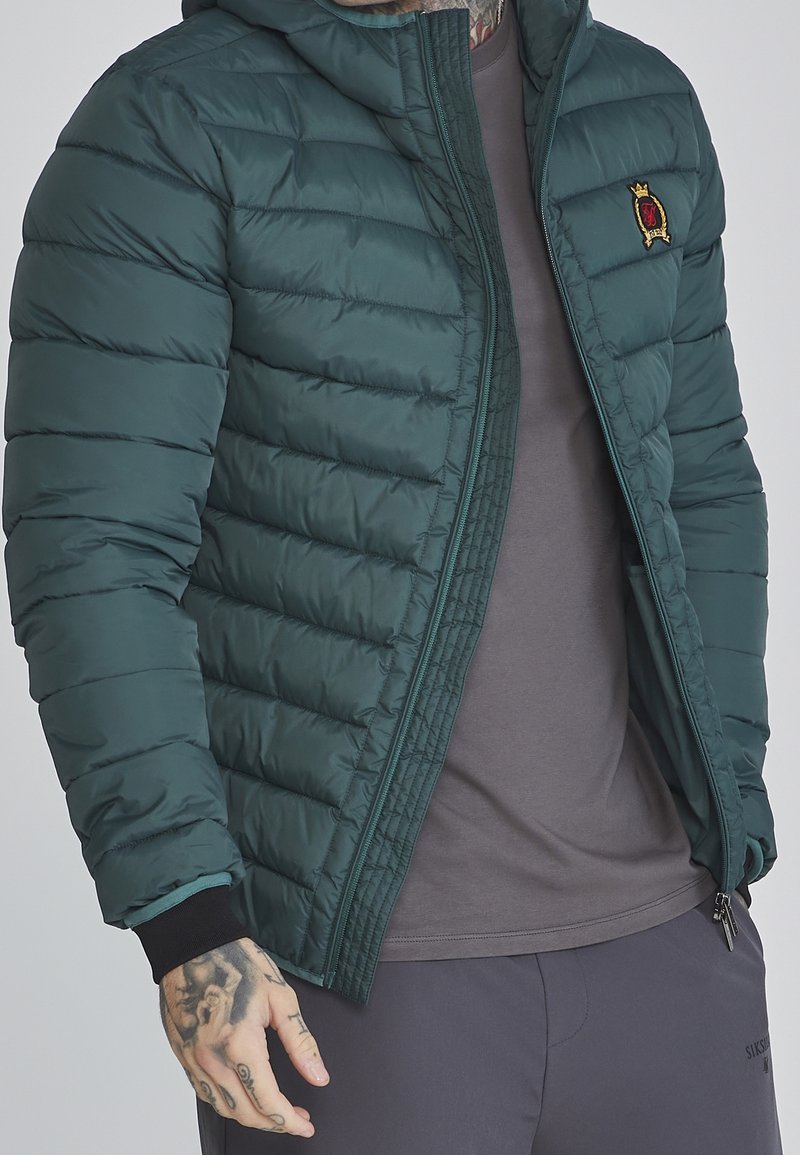 Homme portant une veste matelassée vert foncé par-dessus un t-shirt gris et un pantalon foncé, avec des tatouages visibles sur la main gauche et le cou.
