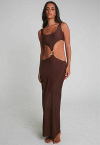 Een model staat blootvoets, gekleed in een slank, donkerbruin jurk met een uitsnijdingen en een zilveren lus rond de taille. De achtergrond is neutraal.