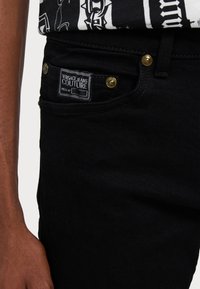 Črne jeans hlače iz denima z zlatim okovjem, ki imajo sprednji žep in oznako Versace Jeans Couture na pasu.