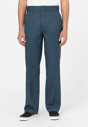 Hombre con pantalones rectos de color azul oscuro y zapatillas negras sin cordones, de pie contra un fondo claro y liso.