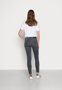 Woman with long brown hair wearing a white t-shirt, dark gray skinny jeans, and white sneakers standing with her back to the camera.