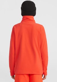 Orange fleece-tröja med hög krage, långa ärmar och mjuk textur. Bärs tillsammans med en svart stickad mössa. Enkelt design, inga synliga logotyper.