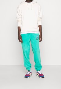 Ljusbeige hoodie kombinerad med tealfärgade sammetsbyxor med tryckt grafik och svart text, kompletterad med flerfärgade sportskor.