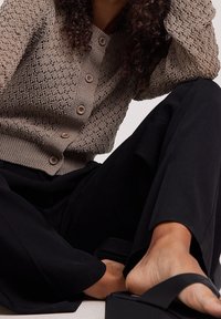 Vaaleanbeige neulottu neuletakki, jossa on teksturoitu kuvio ja napitus edessä. Yhdistetty löysiin mustiin housuihin ja mustiin korokepolkujen sandaaleihin.